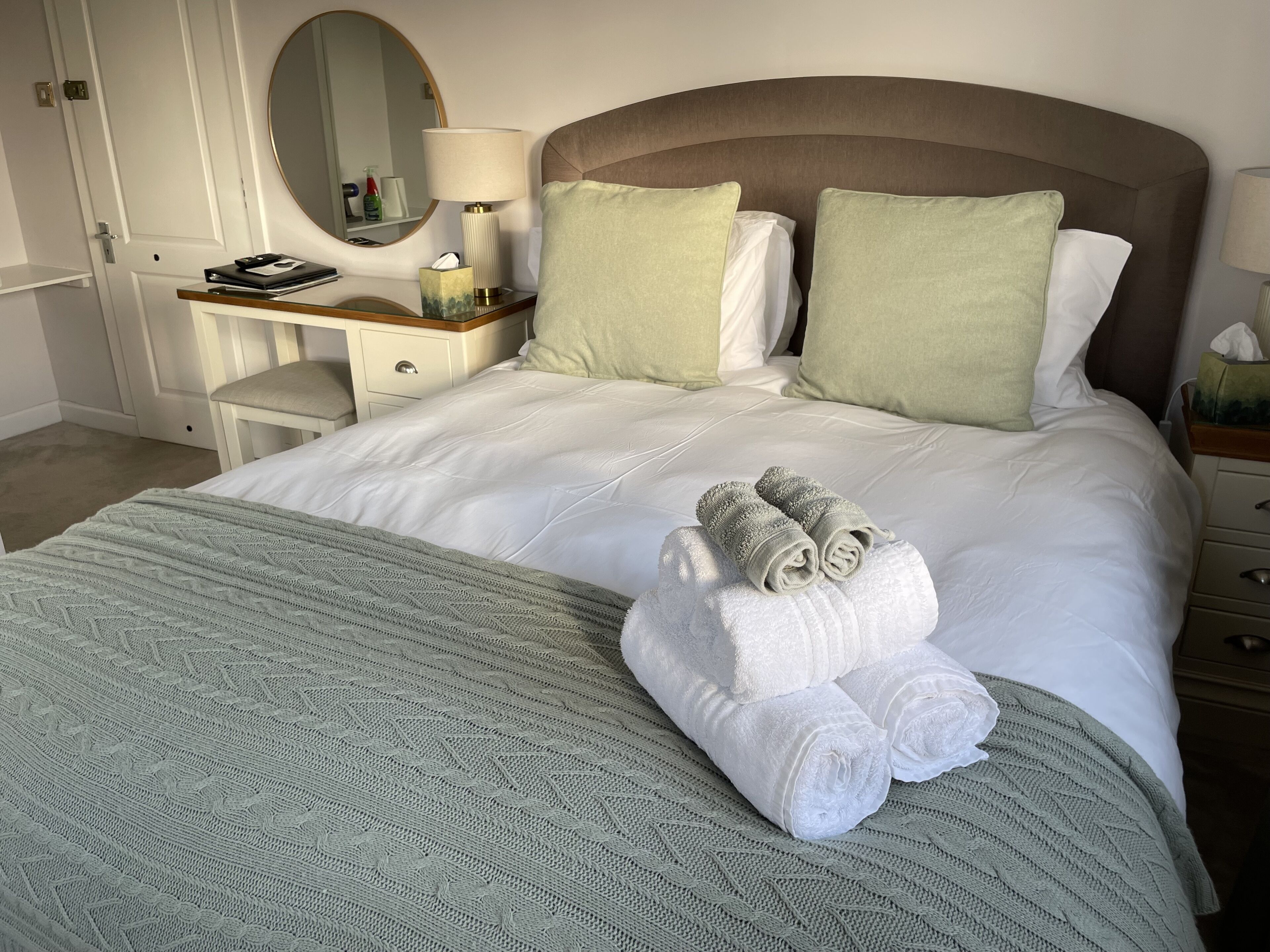 A neatly made bed with white linens and two green pillows, a bedside table with a lamp, a mirror, and a small shelf with toiletries.