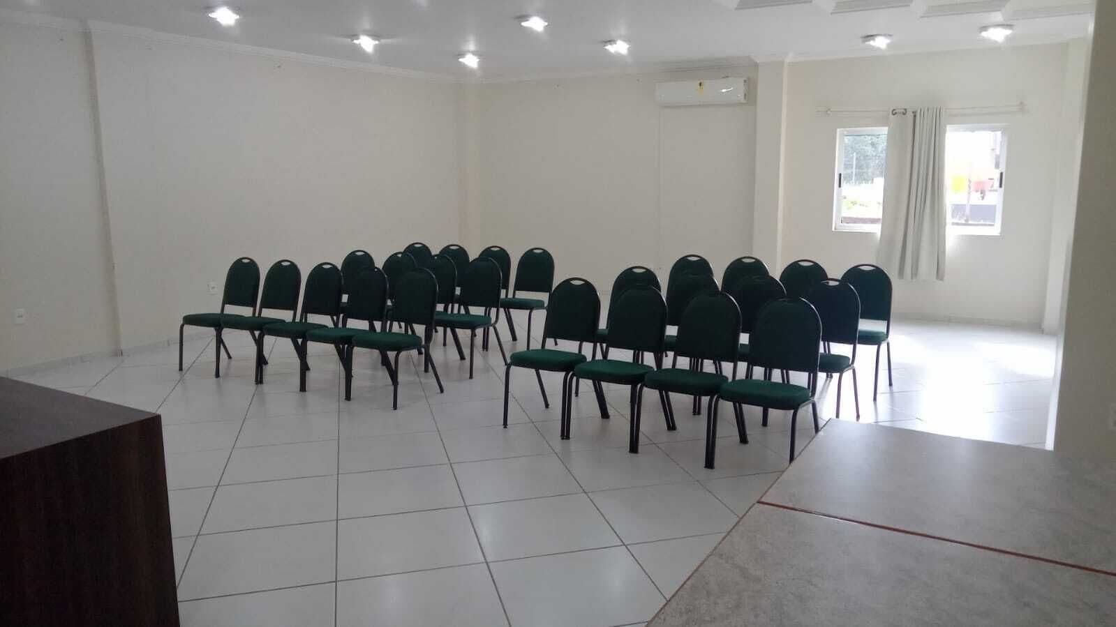 A conference room with rows of green chairs, a large table, white tiled floor, and a window with curtains.