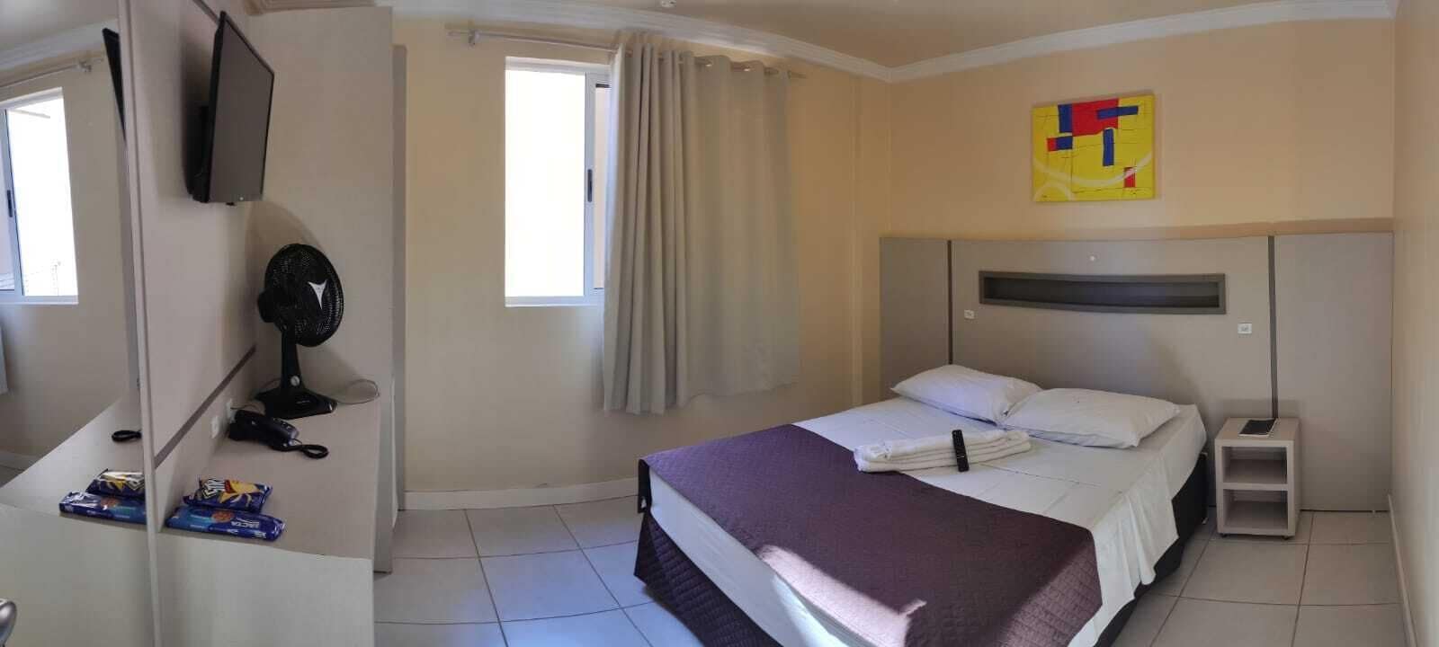 Habitación de hotel con cama, escritorio, computadora, televisor y ventana con cortinas.