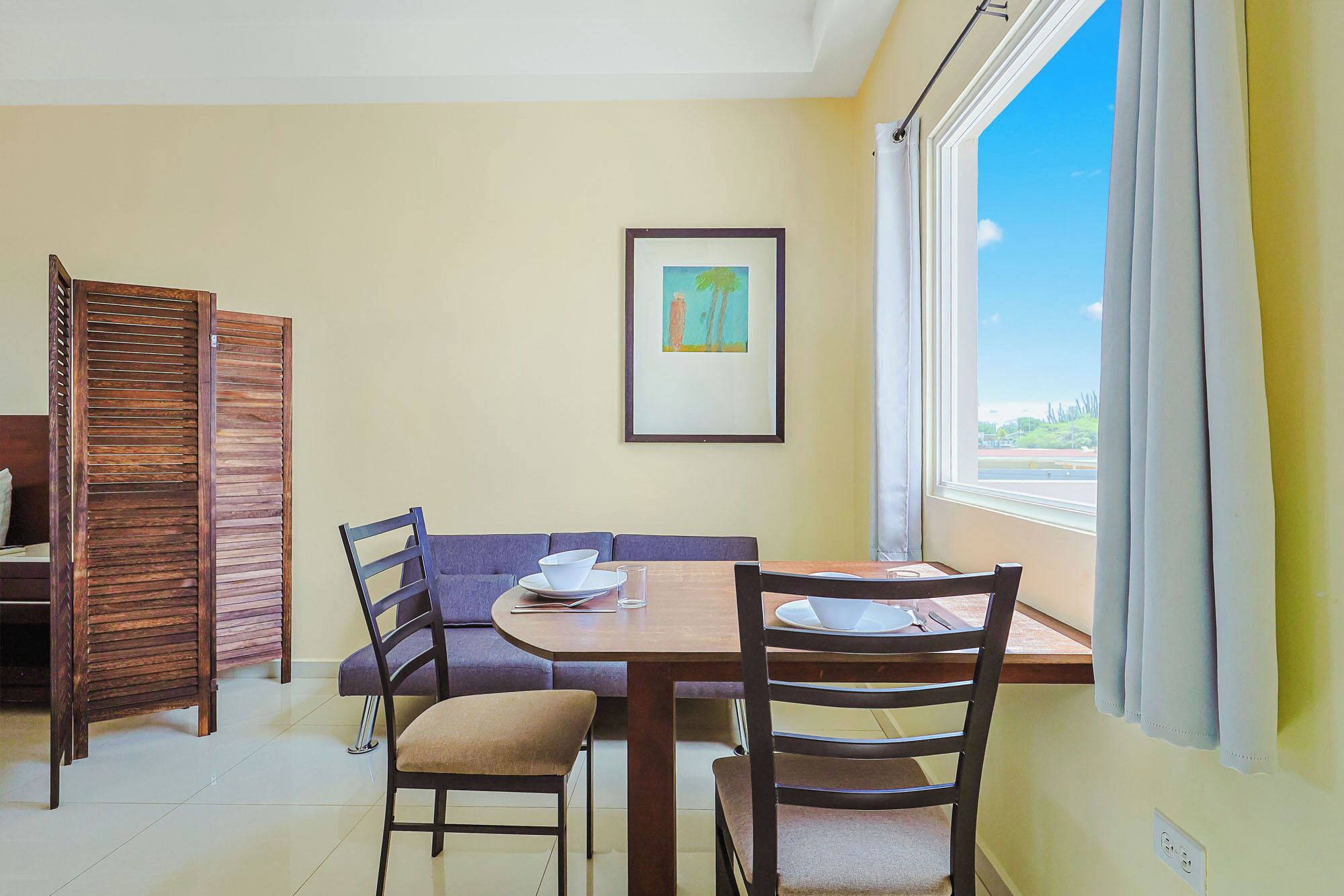 A dining area with a table, chairs, a sofa, a framed picture on the wall, and a window with curtains.
