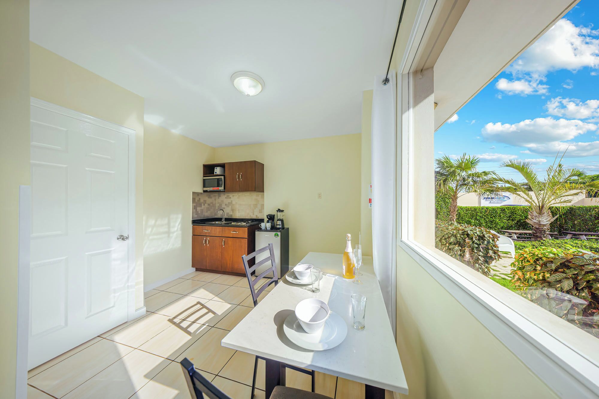 A compact kitchen with a dining area, a white door, and a view of a tropical garden through a window.