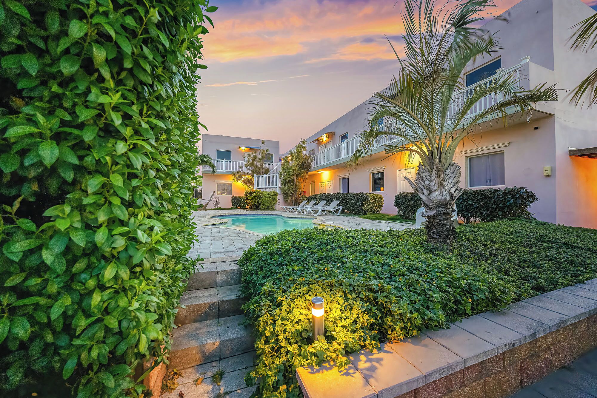 A pool area with lounge chairs, a palm tree, and a staircase leading to a building with balconies.