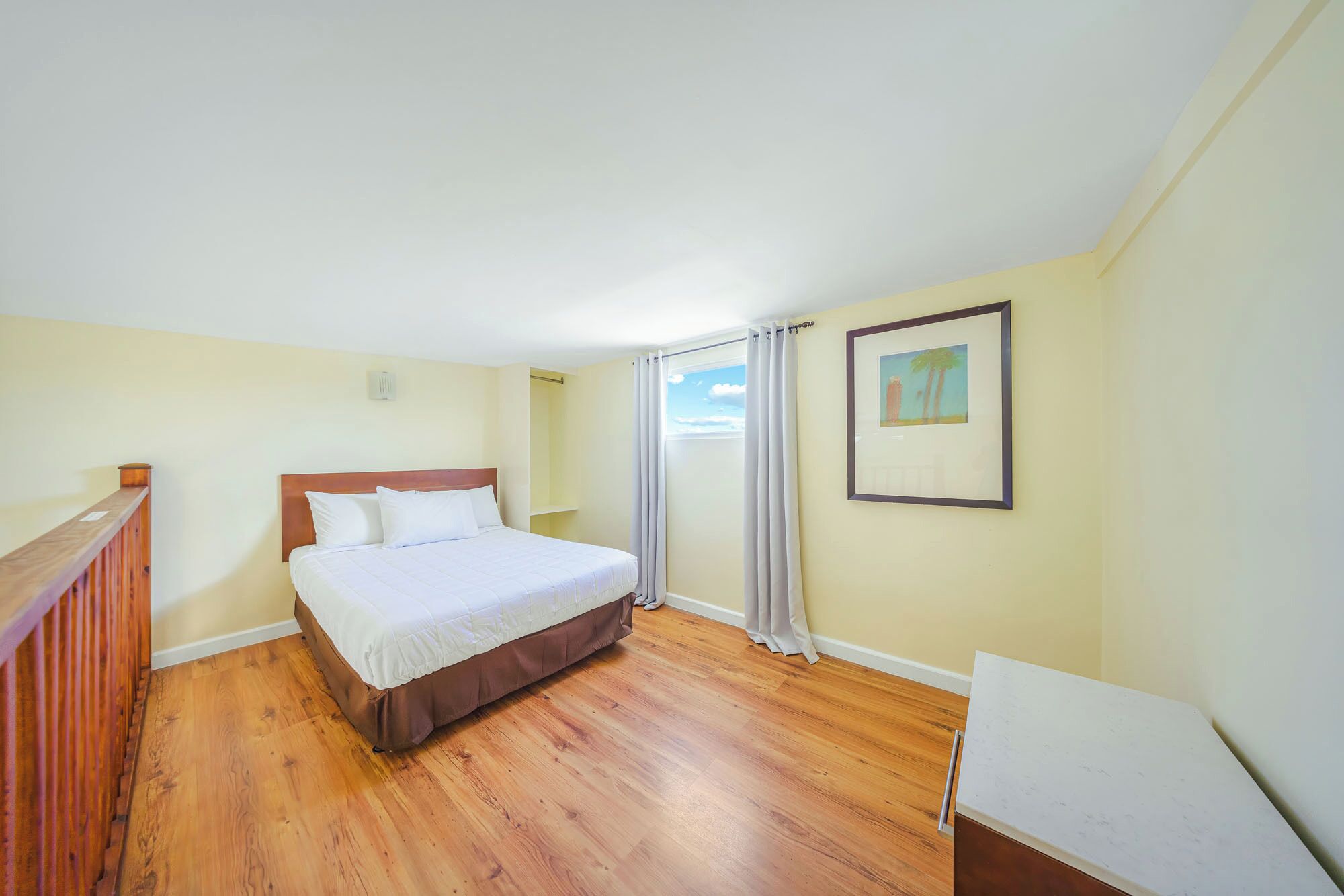 A bedroom with a wooden floor, a bed with white bedding, a wooden headboard, a window with curtains, and a framed picture on the wall.