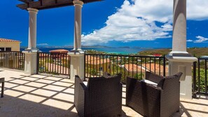 A balcony with wicker furniture, white columns, and a view of the sea and buildings. - The Hills, St John (St. John)