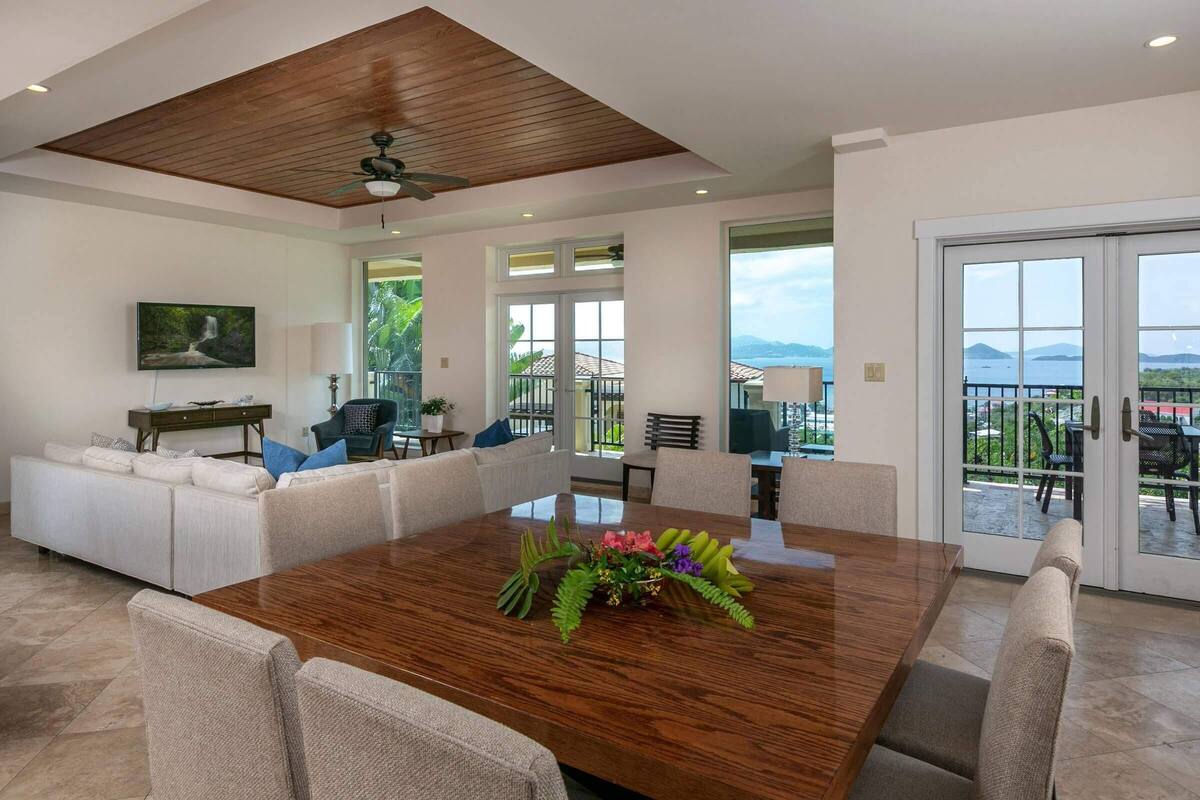 A spacious living area with a wooden dining table, comfortable seating, a ceiling fan, and a view of the outdoors.