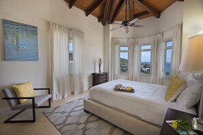 A bedroom with a large bed, a chair, a desk with a lamp, and a view of the sea through windows. - The Hills, St John (St. John)
