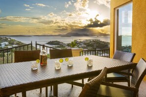 A dining table with chairs, a bottle, and glasses on a balcony overlooking a scenic view of a body of water and distant land. - The Hills, St John (St. John)