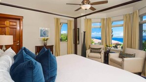 A hotel room with a bed, two chairs, a desk, a ceiling fan, and large windows with a view of the ocean. - The Hills, St John (St. John)