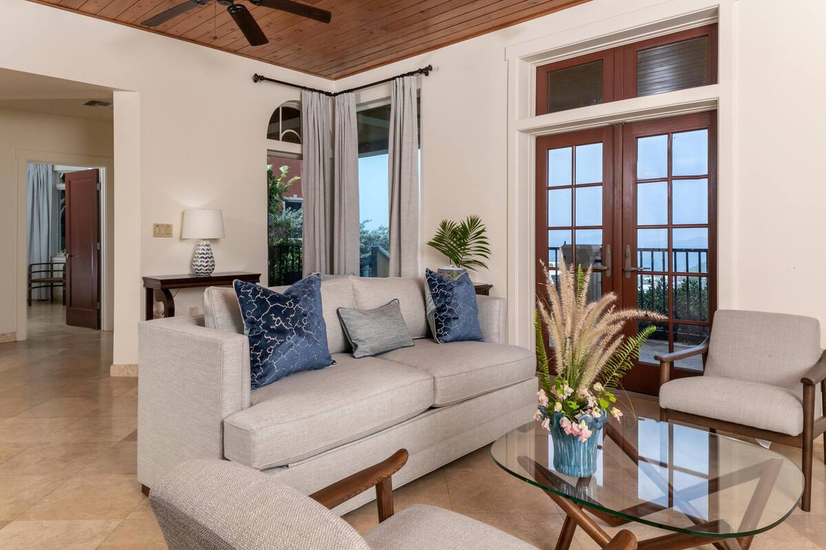 A living room with a beige sofa, a glass coffee table, and a chair. There is a door with a view of the outside.