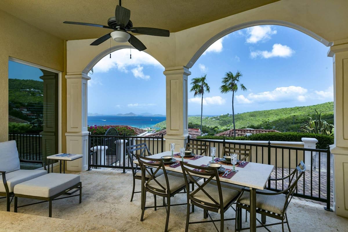 A covered outdoor dining area with a table set for four, overlooking a scenic view of the ocean and hills.