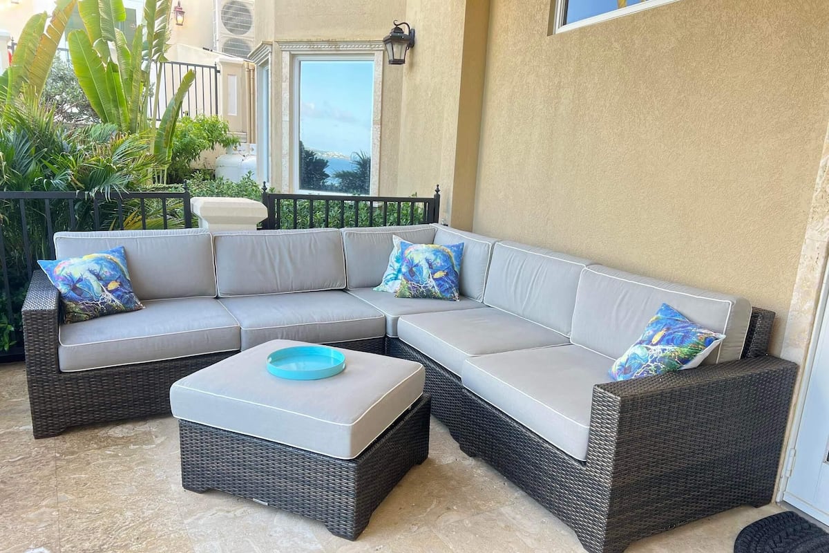 A sectional sofa with cushions, a small ottoman, and a wicker coffee table on a patio with a view of greenery and a building.