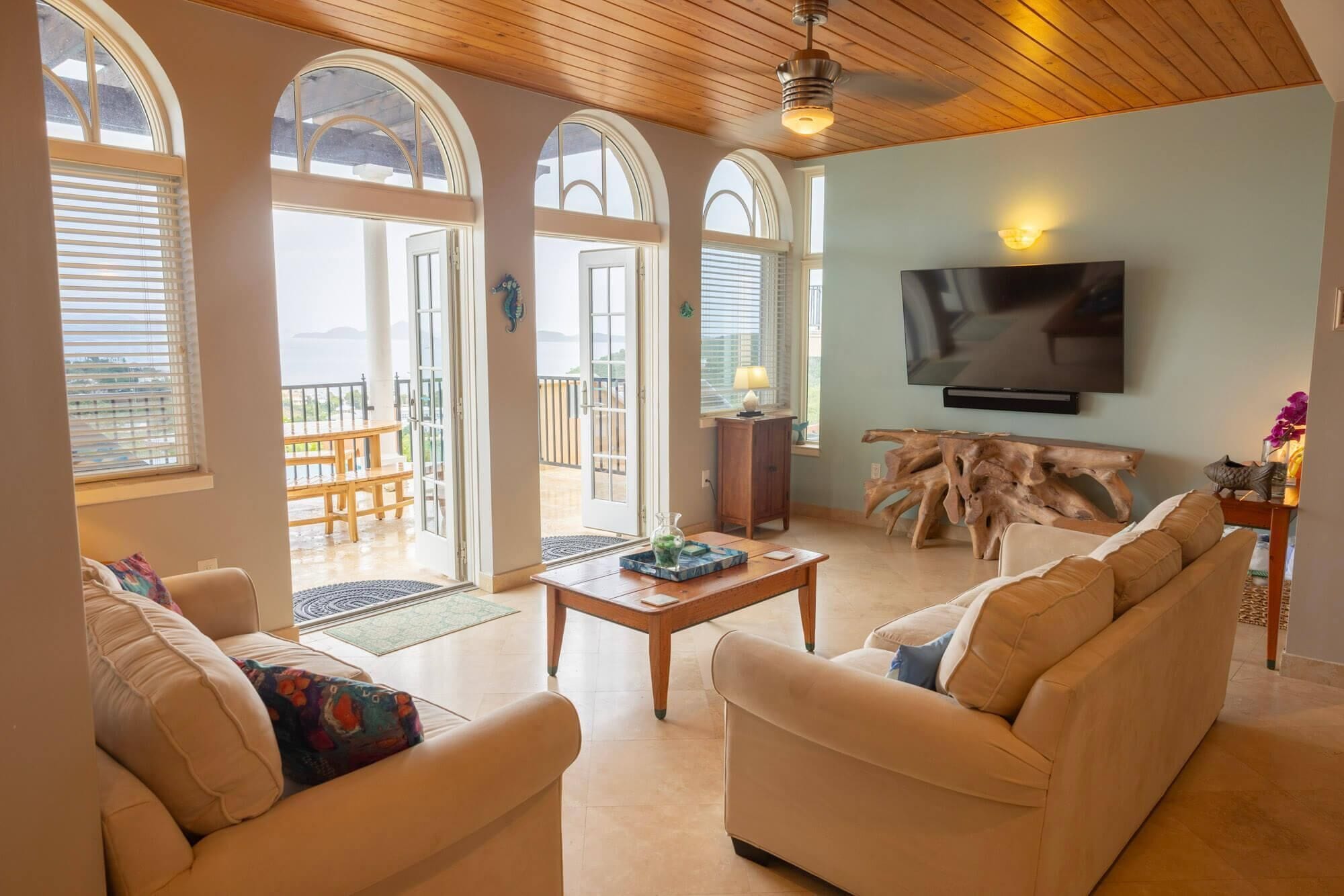 Ein Wohnzimmer mit Sofa, Couchtisch und Fernseher. Es gibt Bogenfenster und einen Balkon mit Tisch und Stühlen.