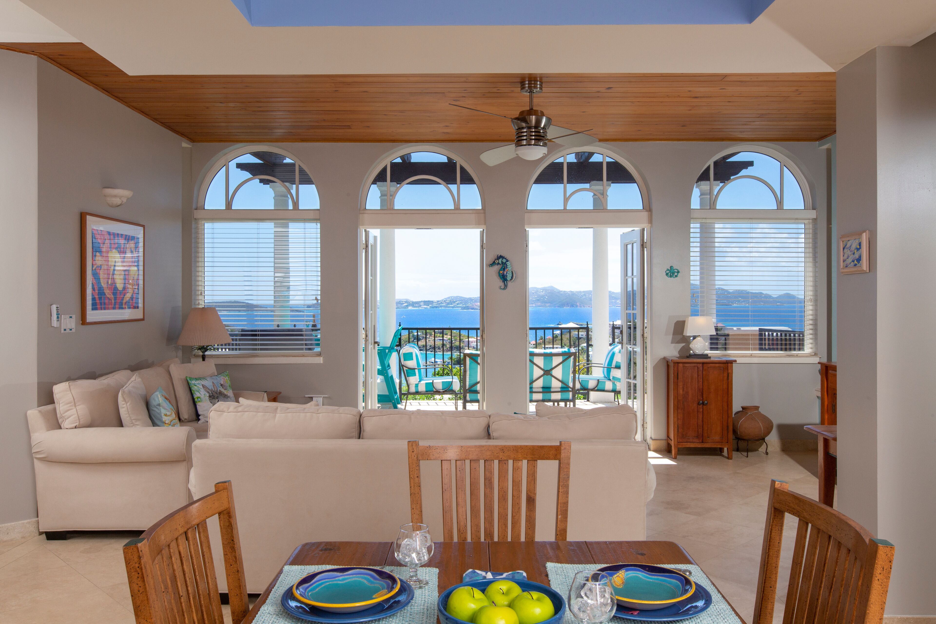 Amplia sala de estar con techo de madera, una mesa de comedor con platos y manzanas, un sofá y vistas al mar a través de amplias ventanas.