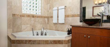 A bathroom with a large bathtub, a wooden vanity, a mirror, and a window with a decorative view.