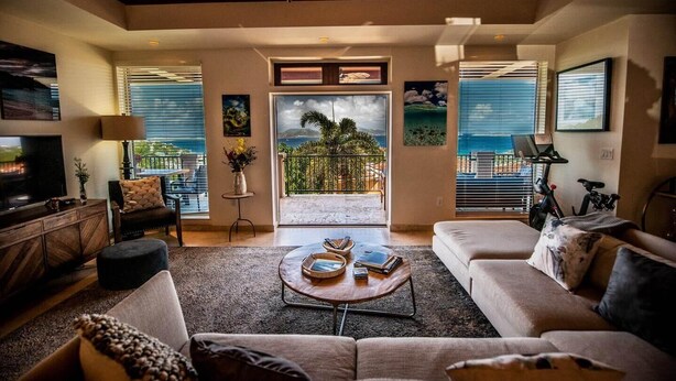 A spacious living room with a large sofa, a coffee table, and a television. There is a balcony with a view of the ocean. - The Hills, St John (St. John)