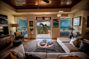 A spacious living room with a large sofa, a coffee table, and a television. There is a balcony with a view of the ocean. - The Hills, St John (St. John)