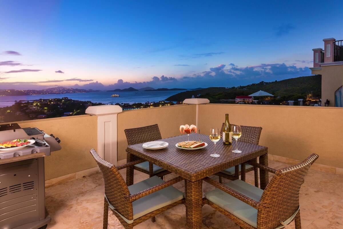 A terrace with a table set for dining, a grill, and a view of the sea and cityscape.
