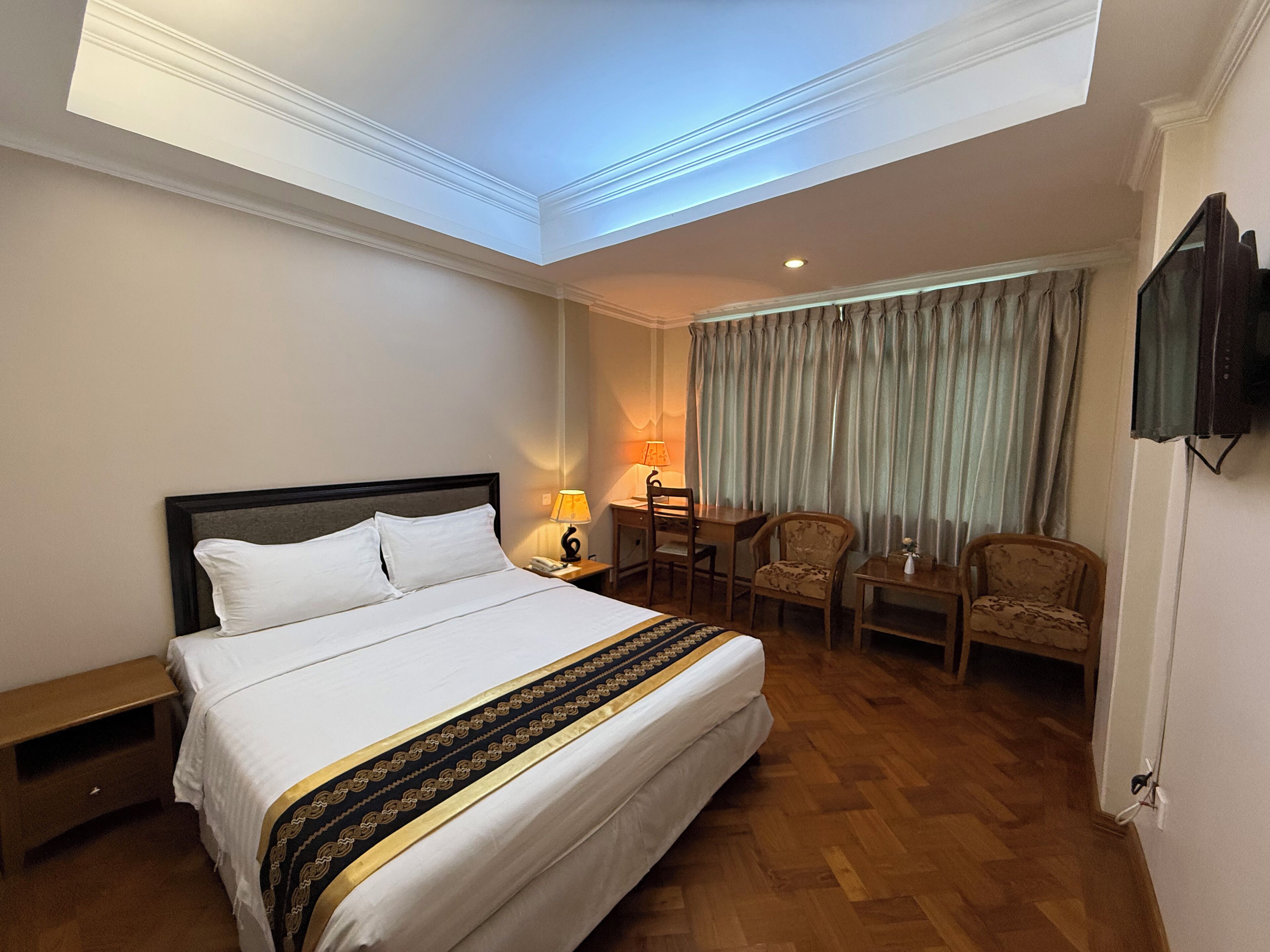 A hotel room with a bed, a desk, two chairs, a television, and a window with curtains.