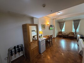A hotel room with a wooden floor, a bed, a desk, a chair, a small refrigerator, a TV, and a window with curtains.