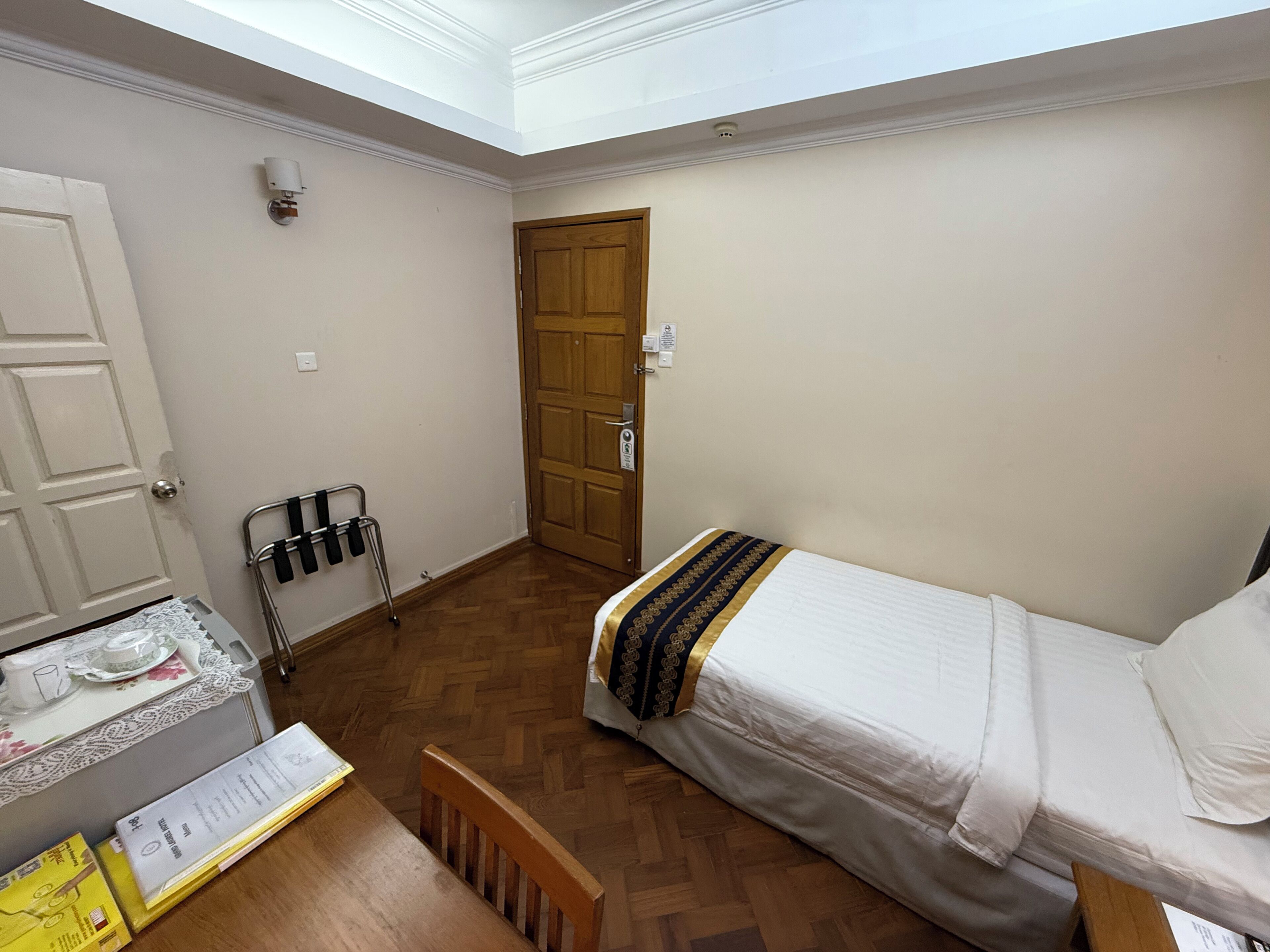 Una habitación pequeña y luminosa con suelo de madera, una cama individual con una colcha estampada, una puerta de madera y una mesa pequeña con mantel de encaje.
