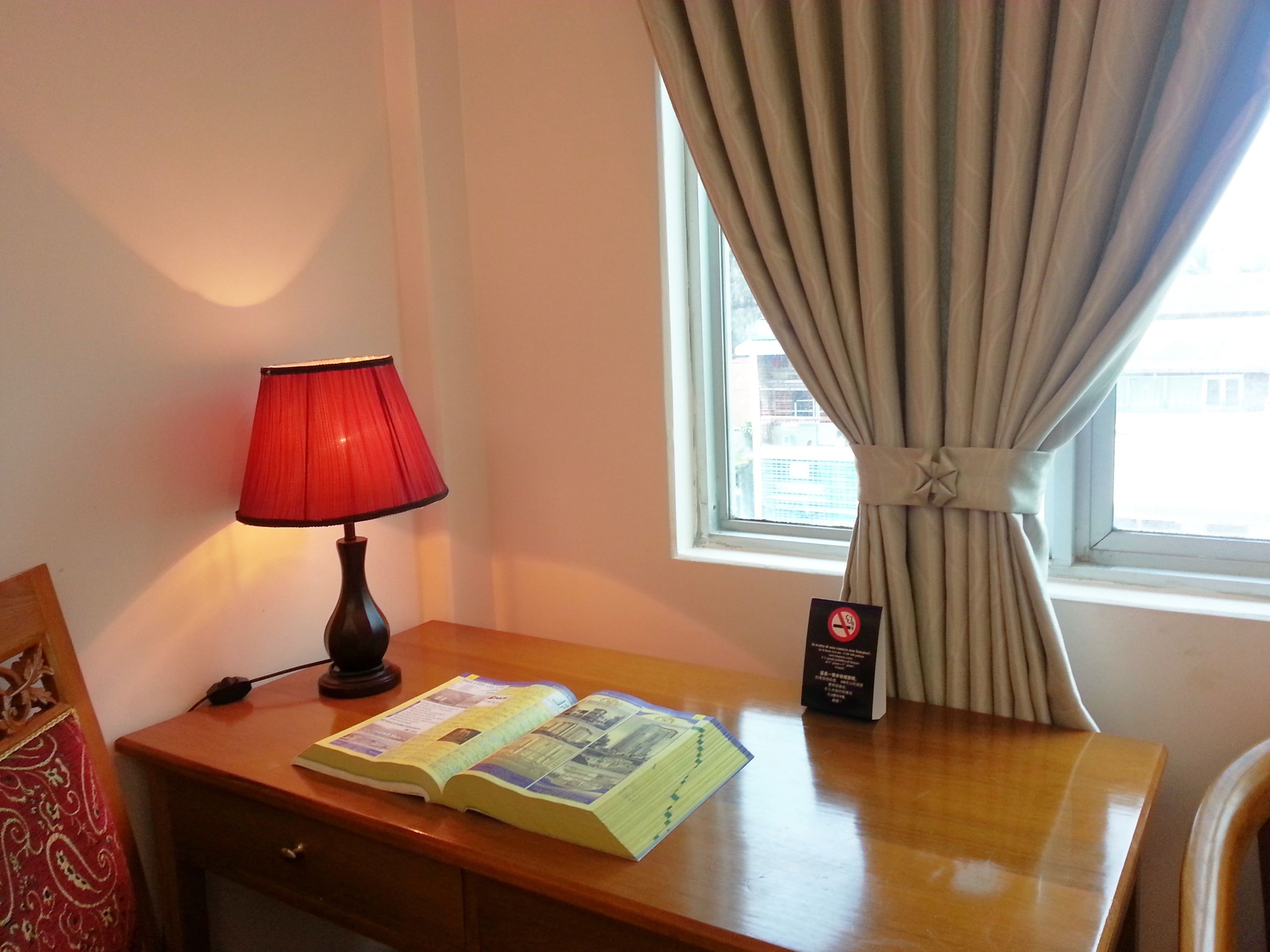 A room with a wooden desk, a red lamp, a book, a no-smoking sign, and a window with curtains.