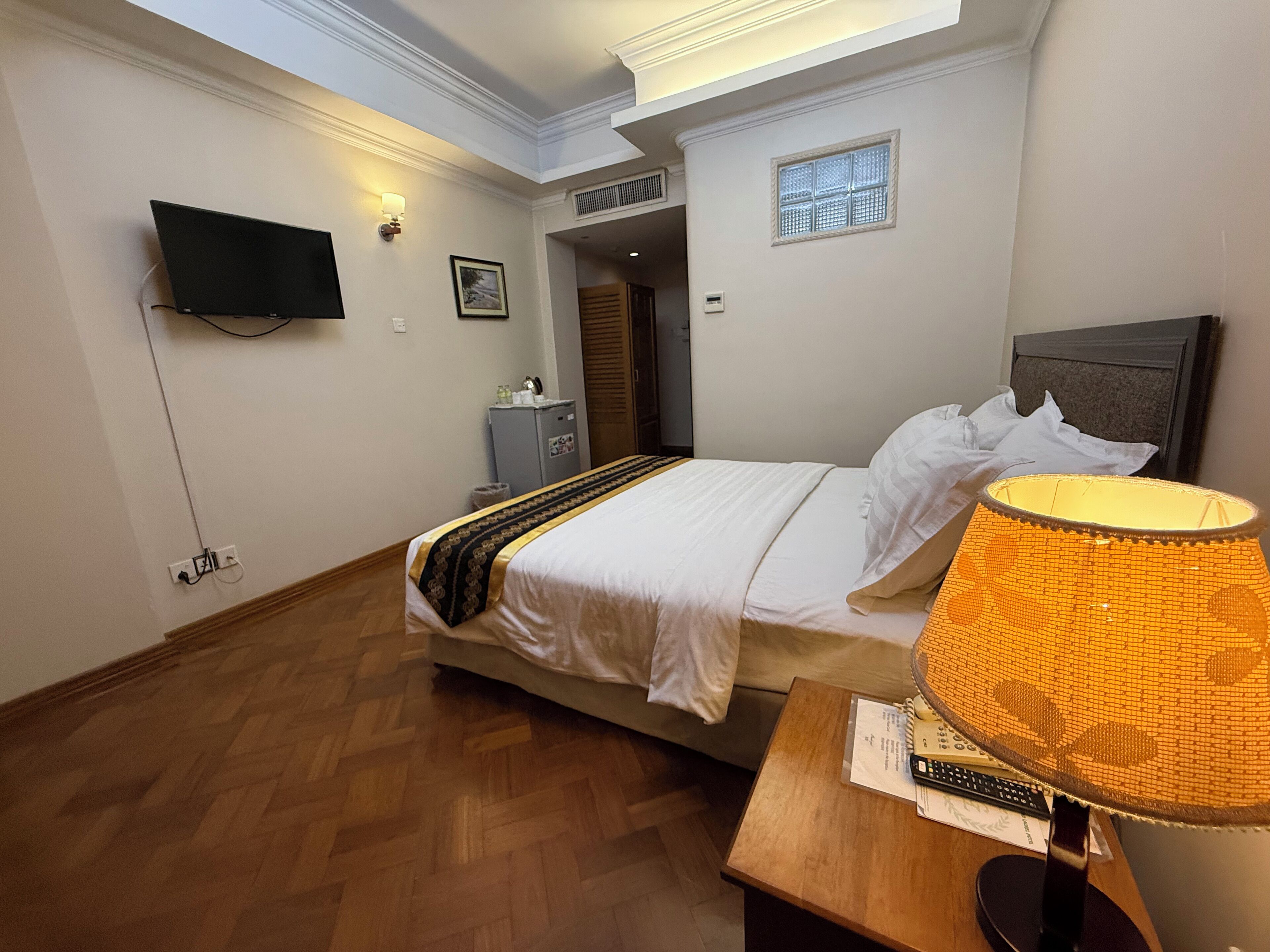 A hotel room with a bed, a desk with a lamp, a television, and a small window.