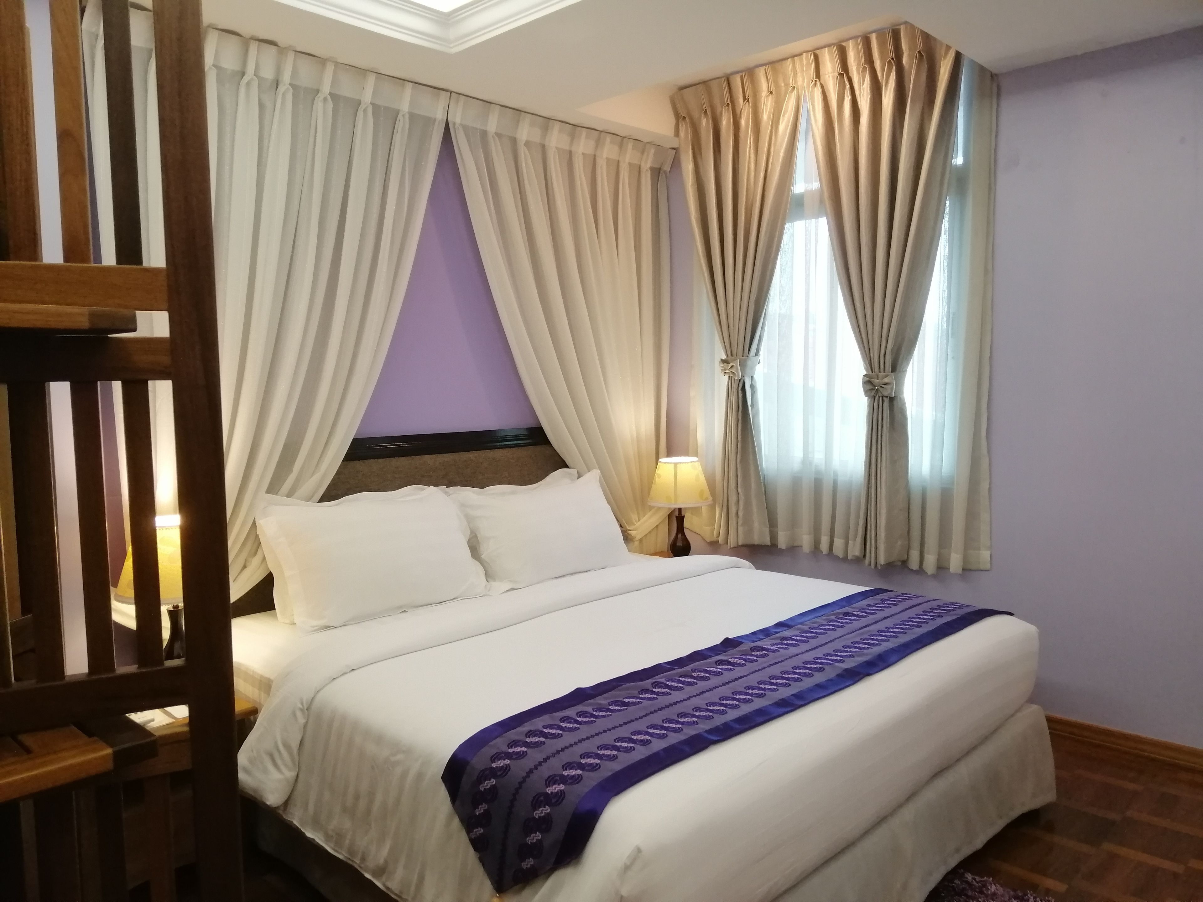 A neatly made bed with white linens and a purple decorative throw, a bedside lamp, and curtains on a window with a view of the outdoors.