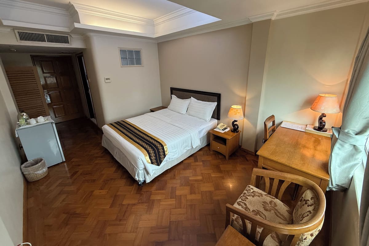 A hotel room with a bed, a desk, a chair, a small table, a lamp, and a refrigerator.