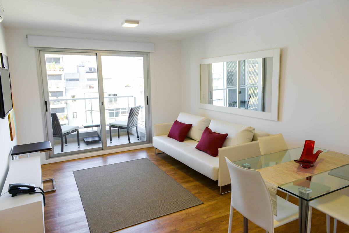 A modern living room with a glass dining table, white sofa with red cushions, and a large sliding glass door leading to a balcony.