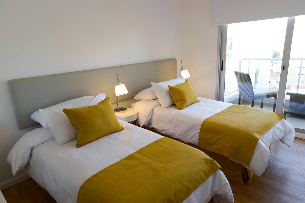 A hotel room with two beds, each with white bedding and yellow accents, a small bedside table with a lamp, and a balcony with a chair.
