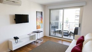 A modern living room with a flat-screen TV, a white sofa with red cushions, a white console table, and a sliding glass door leading to an outdoor balcony. - Mérit Montevideo Apart & Suites (Montevideo)