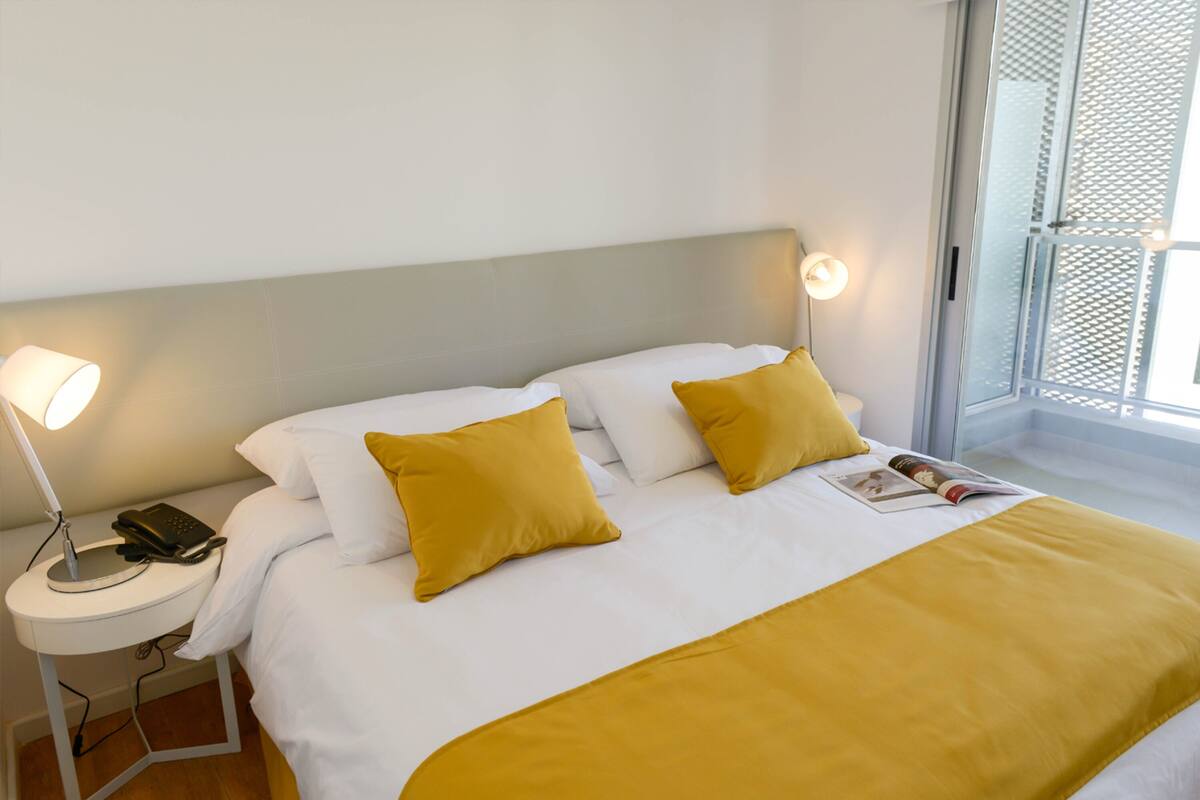 A neatly made bed with white and yellow bedding, a bedside lamp, and a magazine on the bed.