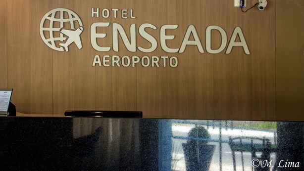 A reception desk with a wooden panel wall displaying the text "Hotel Enseada Aeroporto". - Hotel Enseada Aeroporto (Recife)