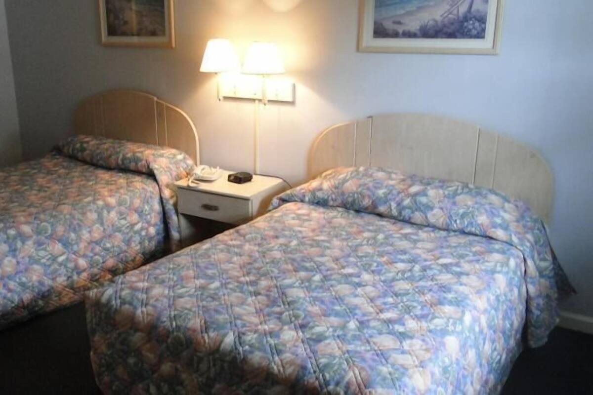 A room with two single beds, a nightstand with a lamp, and a wall-mounted light fixture.