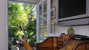 A wooden desk with a chair, a television mounted on the wall, and a window with a view of greenery outside.