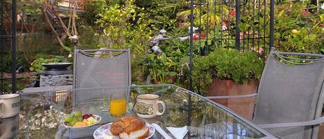 Une table en verre, mise pour le petit-déjeuner, avec un croissant, des fruits et du jus d’orange, entourée de chaises et donnant sur un jardin.