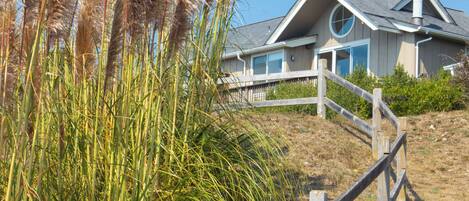 A house with a wooden fence, surrounded by tall grasses and a clear blue sky.