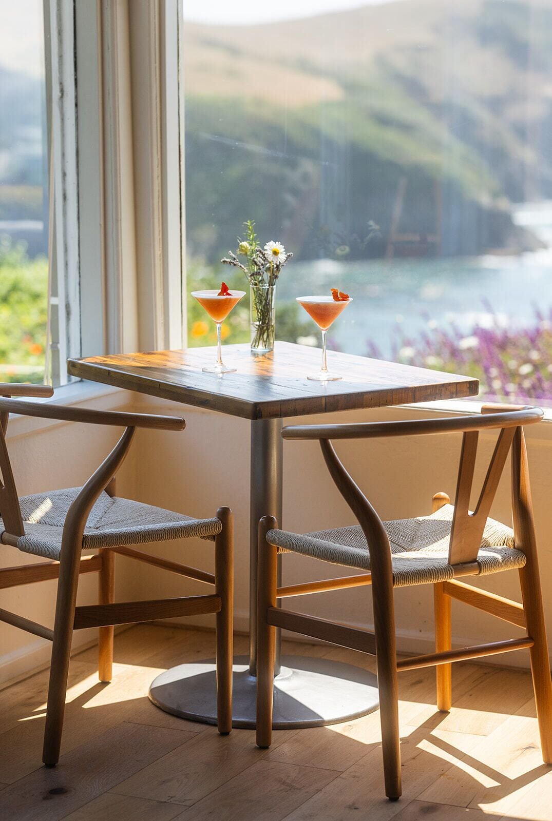 Une table avec deux chaises et deux verres à martini garnis, posée sur un plancher en bois, avec vue sur une étendue d’eau par la fenêtre.