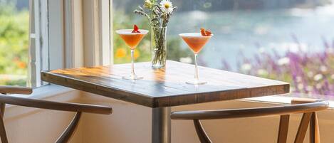 Une table avec deux chaises et deux verres à martini garnis, posée sur un plancher en bois, avec vue sur une étendue d’eau par la fenêtre.