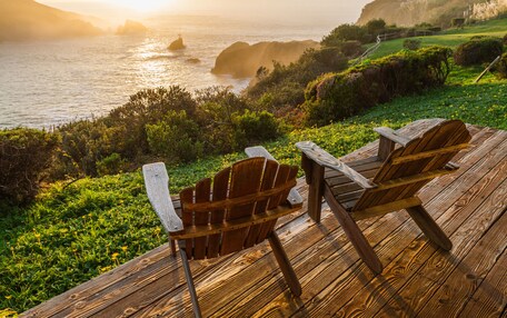 A wooden deck with two Adirondack chairs overlooking a coastal landscape with hills and the ocean.