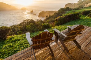 Una terraza de madera con dos sillas Adirondack que dan a un paisaje costero con colinas y el océano.