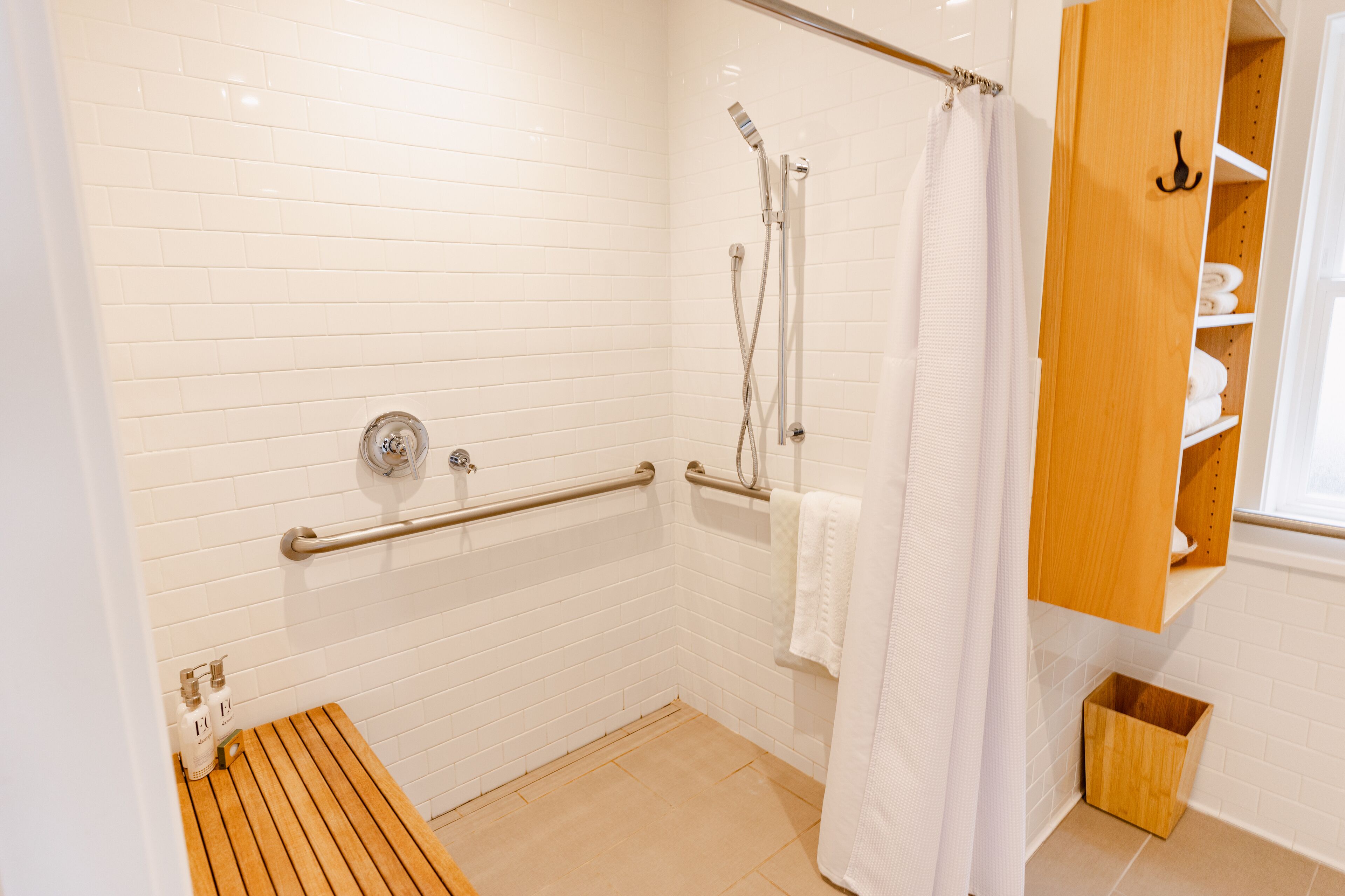 Un baño moderno con paredes de azulejos blancos, un banco de madera, una ducha con puerta de cristal y un armario de madera con estantes.