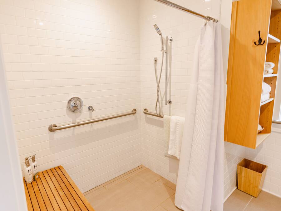 Un baño moderno con paredes de azulejos blancos, un banco de madera, una ducha con puerta de cristal y un armario de madera con estantes.