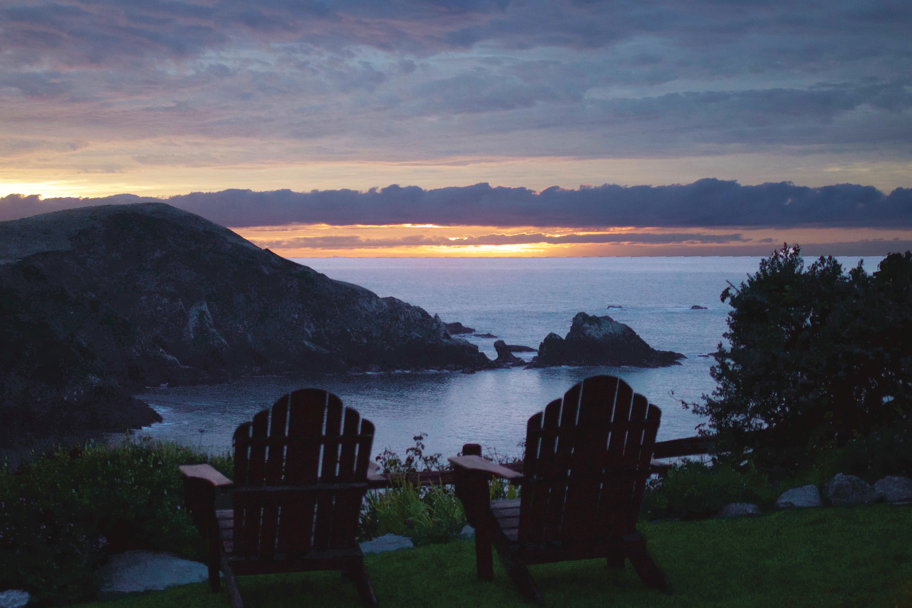 Two Adirondack chairs facing a sunset over a body of water with rocky terrain and hills in the background.