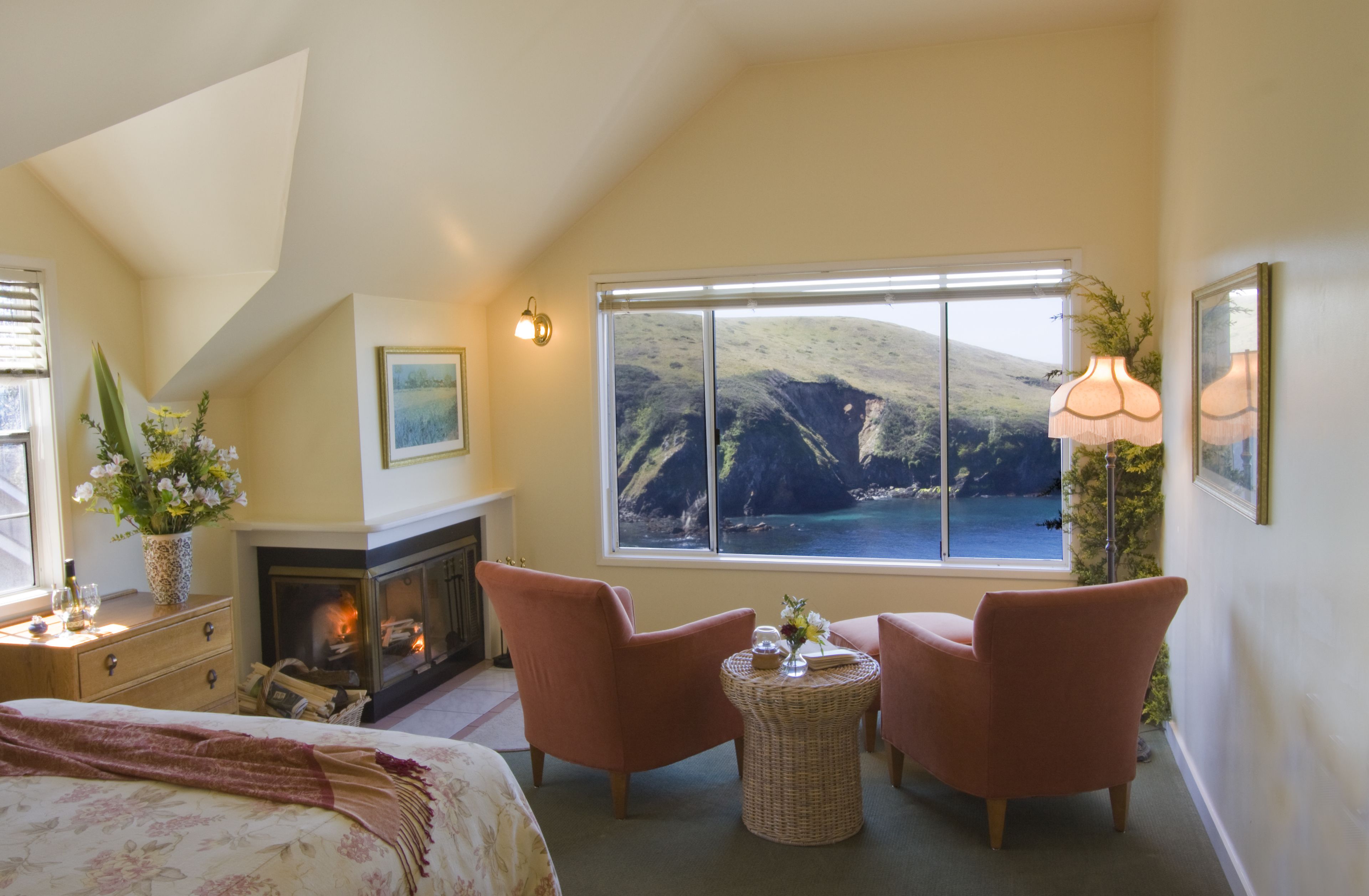 A cozy bedroom with a fireplace, two pink armchairs, a round wicker table, and a large window overlooking a scenic coastal view.