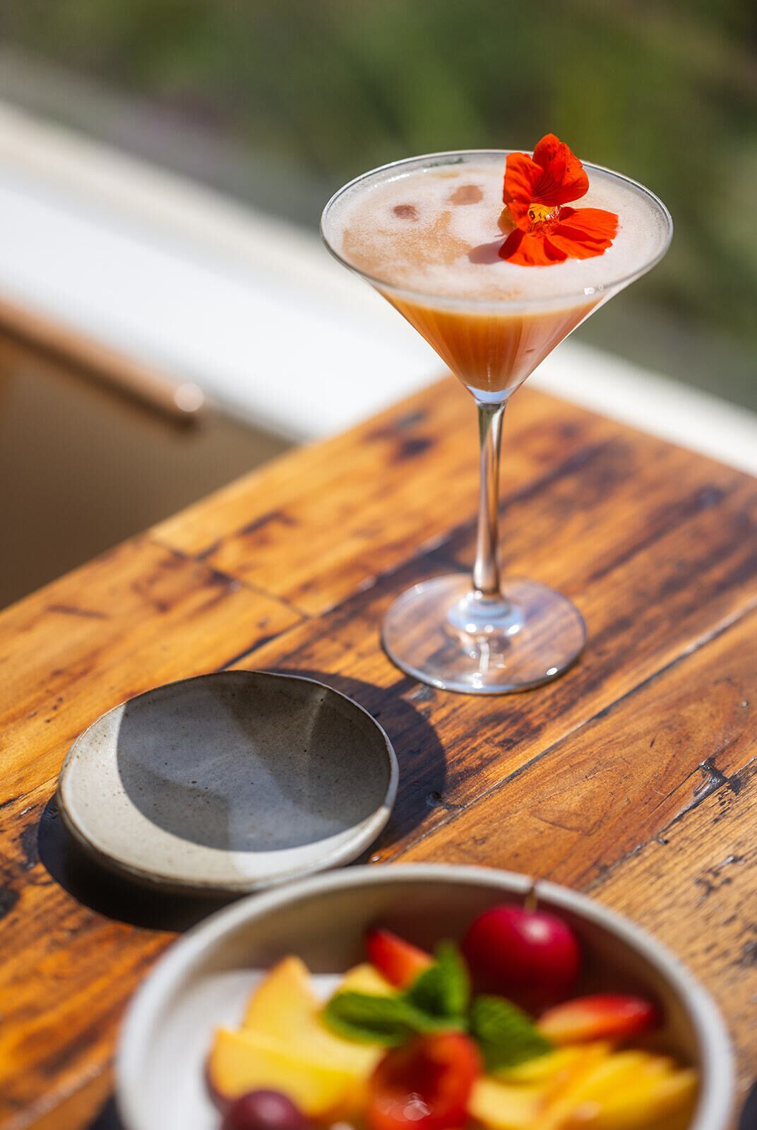 A martini glass with a frothy top and a red flower garnish on a wooden table.