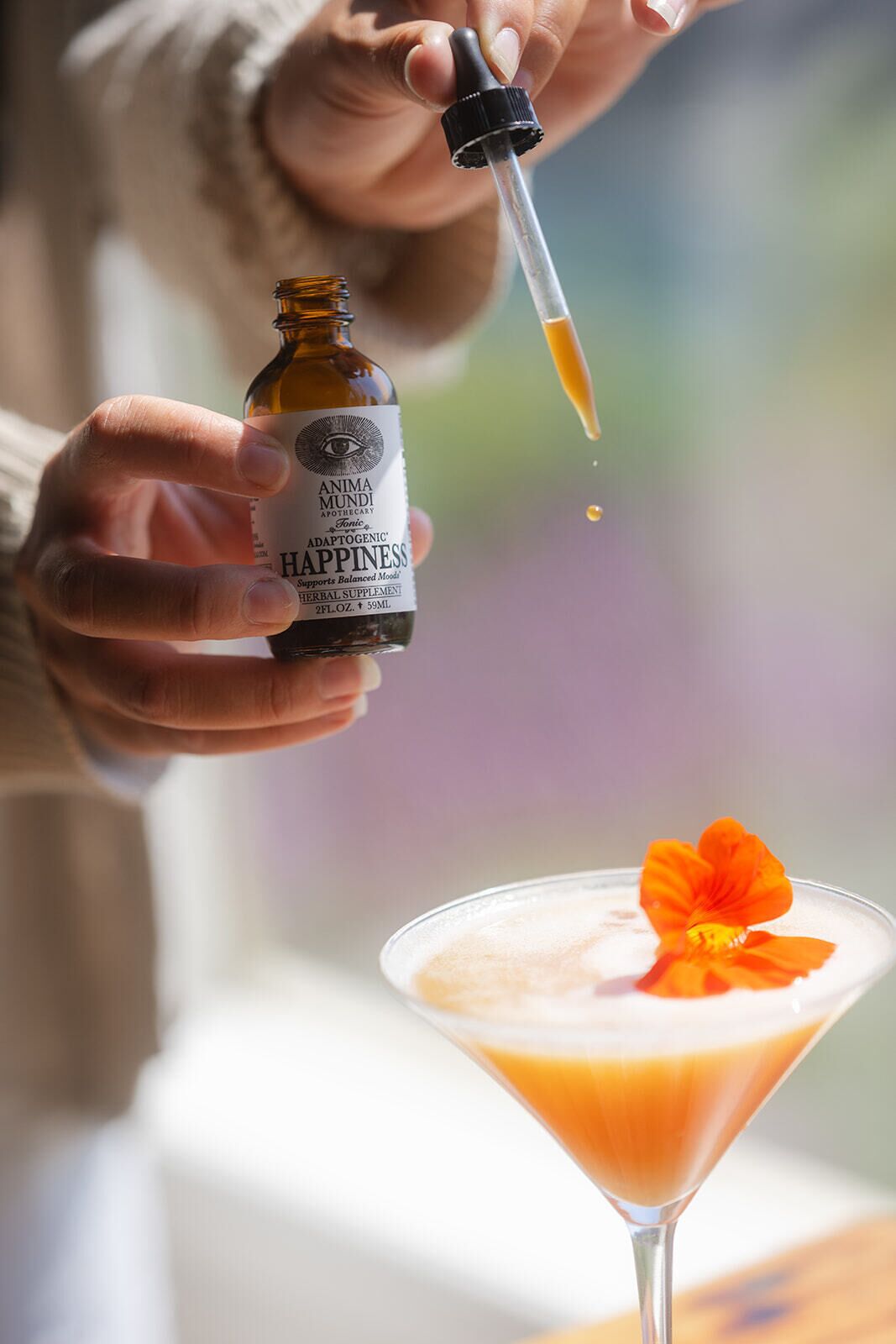 A person holding a bottle labeled 'ANIMA MUNDI' and pouring a liquid into a martini glass with an orange flower garnish.