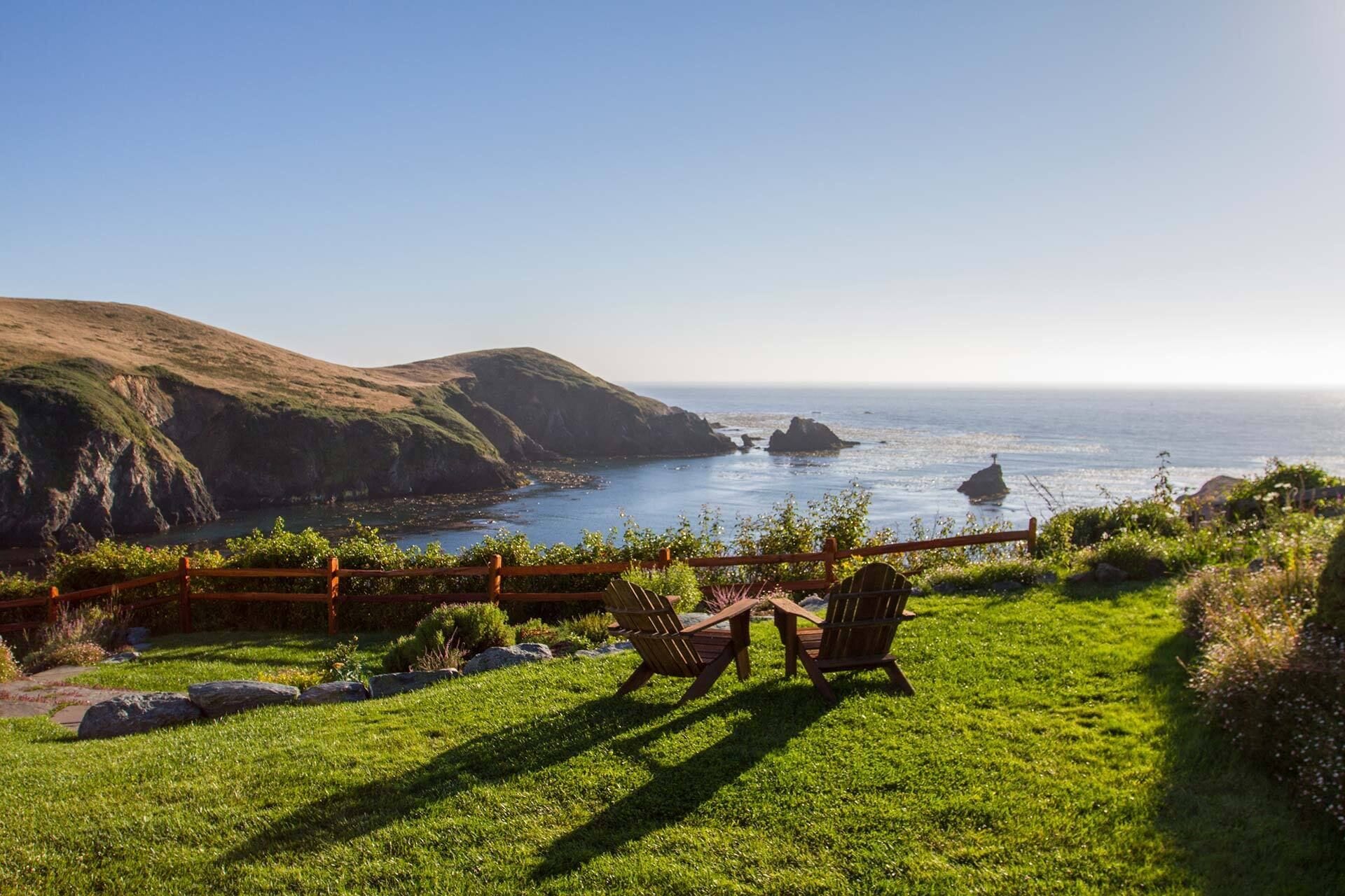 A grassy outdoor area with a wooden fence, two wooden chairs, and a view of the ocean with rocky formations.