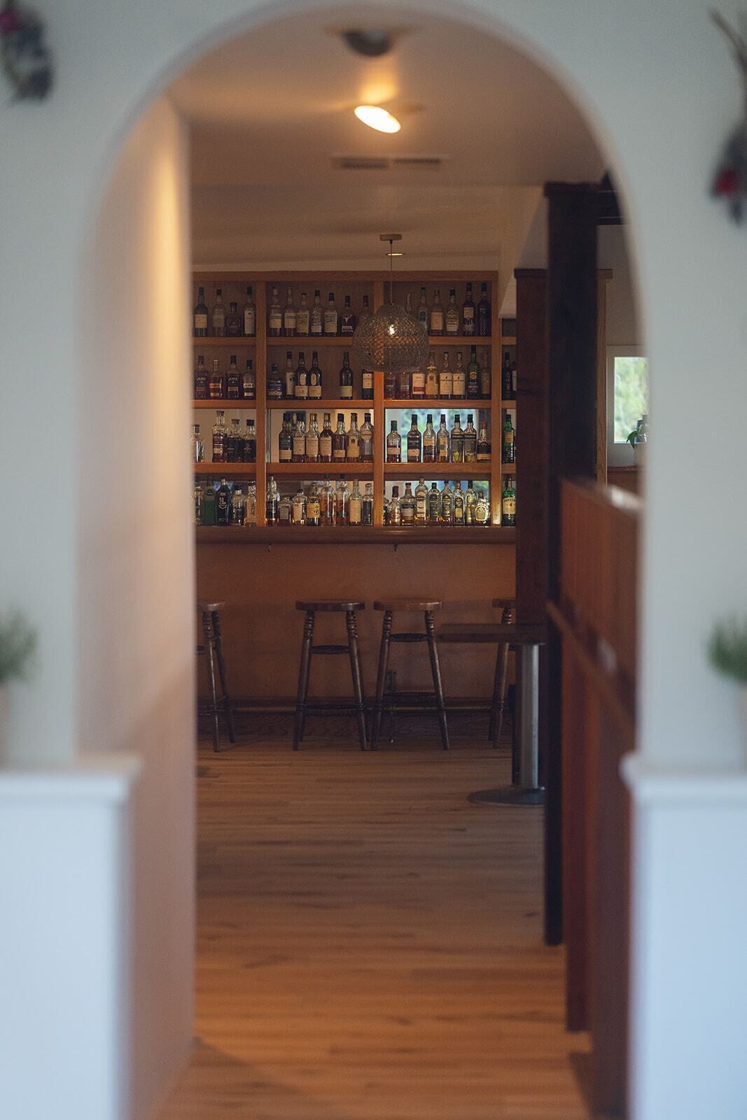Vista interior d'una zona de bar amb prestatges de fusta plens de botelles, una lluminària penjant i un sòl de fusta.