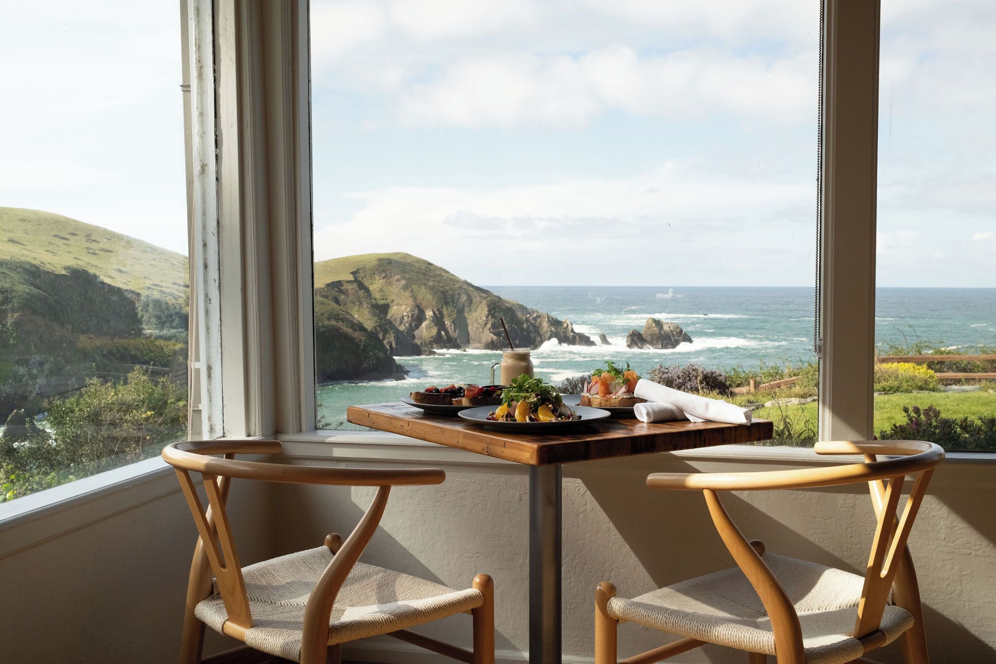 Un balcón con una mesa dispuesta para dos personas, con vistas a un paisaje costero con acantilados y el mar.
